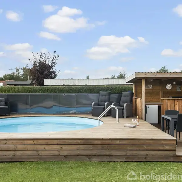 Rund pool på træterrasse med stige. Sofaer og skur ved siden af indeholder spisebord og stole. Græsområde omkranser terrassen under en skyfyldt blå himmel.