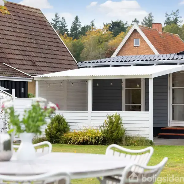 Et hus står i en have med grøn græsplæne, omgivet af flere planter. Foran ses et bord med stole, der skaber en hyggelig udendørs spiseplads.