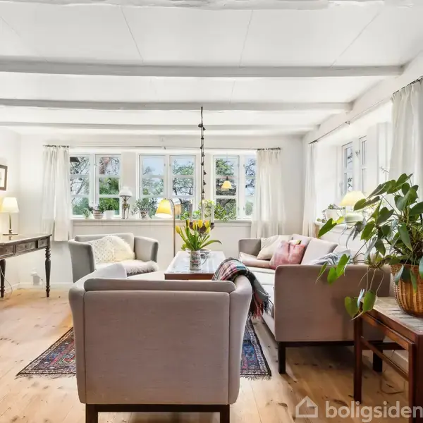 En stue med lysegrå sofaer og en lænestol, centralt placeret omkring et sofabord med blomster. Rummet er lyst med store vinduer og trægulv, omgivet af grønne planter og lamper.