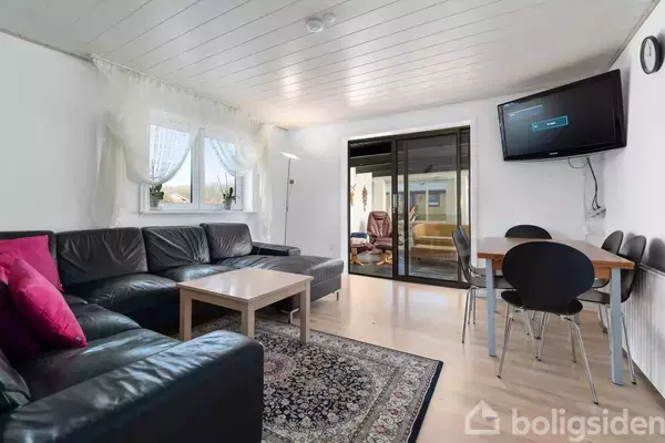 Stue med sort hjørnesofa og træbord på orientalsk tæppe; fladskærm hænger på væggen over et rundt spisebord. Store vinduer med lette gardiner giver naturligt lys.