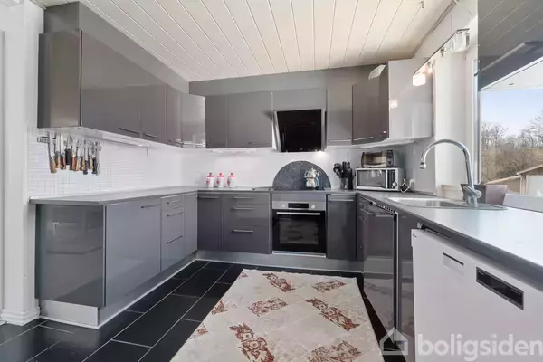 Moderne køkken med grå højglansskabe, indbygget komfur og kogeplade. Krogen har en række knive hængende på væggen. Et lyst tæppe ligger på det sorte klinkegulv.