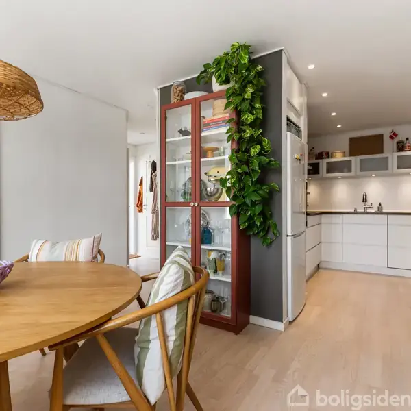 Et rundt træbord med farverig skål står i lys spisestue. Ved siden af står et vitrineskab med grøn plante. Bagved ses et moderne køkken med hvide skabe og indbygget lys.