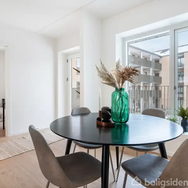 Et rundt spisebord med fire stole står i et lyst rum. En grøn vase med tørrede blomster pryder bordet. Udsigt til altan og anden bygning ses gennem store vinduer.