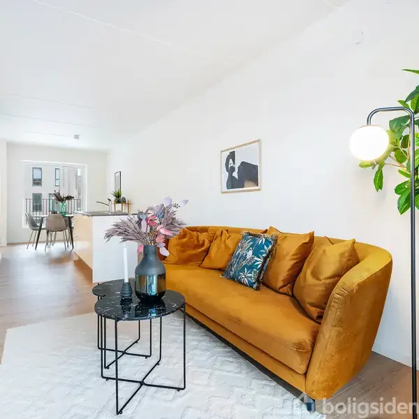 En gul sofa står på et lyst tæppe ved siden af et rundt sofabord med vase i en moderne stue. En grøn plante og vægbillede er baggrunden.