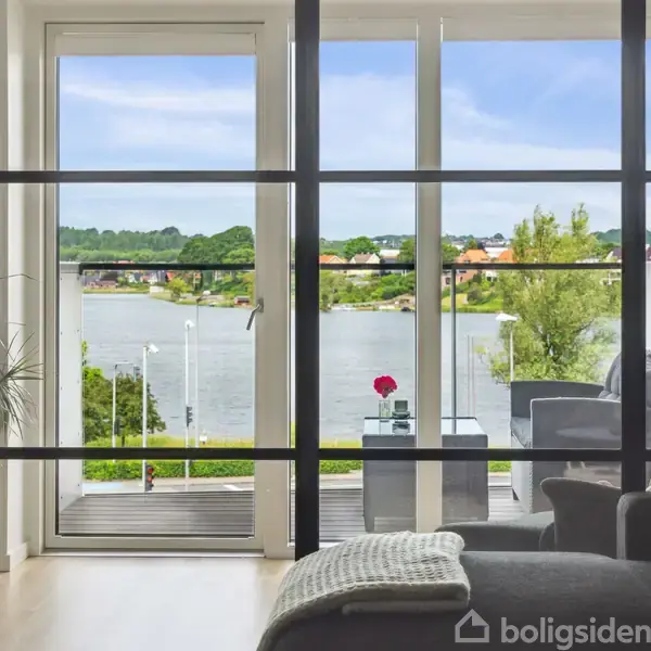 Et lyst værelse med glasdøre åbner ud til en terrasse med udsigt over en sø og grønne områder. Indenfor ses en plante og en lænestol.