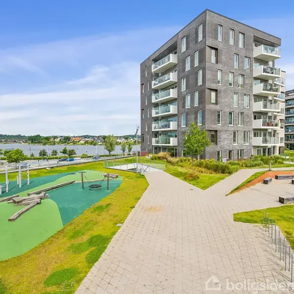 Et moderne lejlighedskompleks med altaner står ved siden af en legeplads med gynger og grønne områder. I baggrunden ses vand og en park.