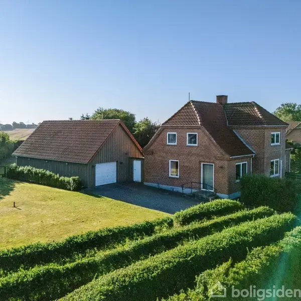 Rødt murstenshus med rødt tag står stille ved siden af en garagebygning. Omkring er der en velplejet have med hække. Landskab med marker i baggrunden og blå himmel.