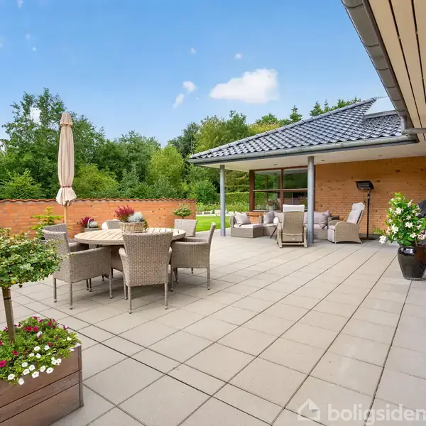 Terrasse med spisebord og stole, omgivet af blomsterkrukker. En parasol står ved bordet. Til højre er der udendørs lounge-møbler. Rød murstensvæg omkranser området, der er omgivet af træer.