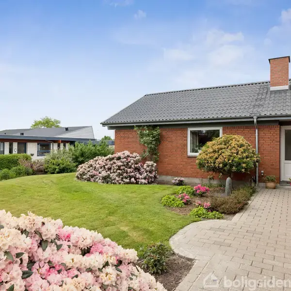 En murstensbungalow med mørkt tag står ved siden af en brostensbelagt sti. Farverige buske og blomsterbed omgiver huset i en velholdt have under en klar, blå himmel.