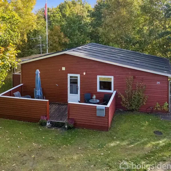 Rødt hus med sort tag står omgivet af grønne træer. En lille terrasse har parasol og møbler. Huset har nummeret 26 ved siden af døren. Græsplæne omkranser huset.