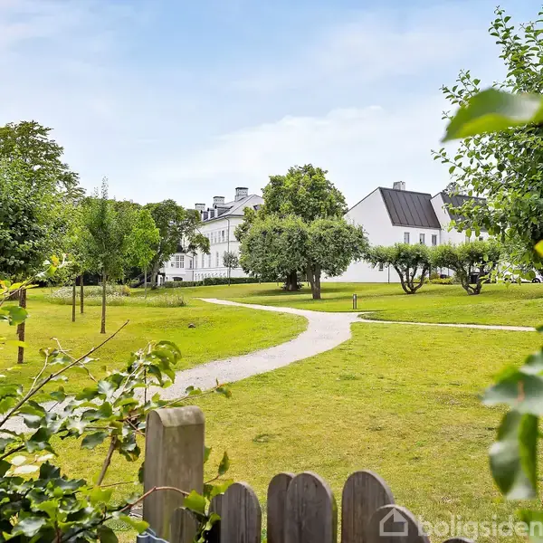 En hvid bygning står for enden af en snoet sti omgivet af en grøn park med træer og græs. Himmel og skyer ses i baggrunden.