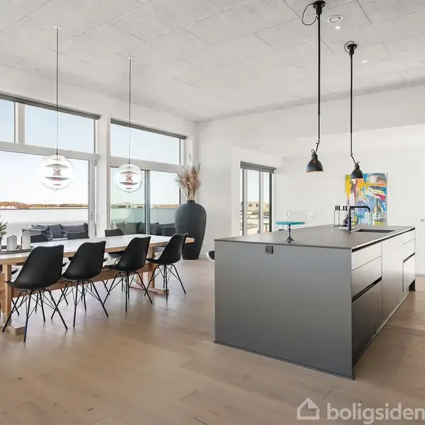 Køkken-alrum med moderne køkkenø, hængende lamper, og spisebord med sorte stole. Store vinduer åbner til udsigt over terrasse med plantekrukker og havet i baggrunden.