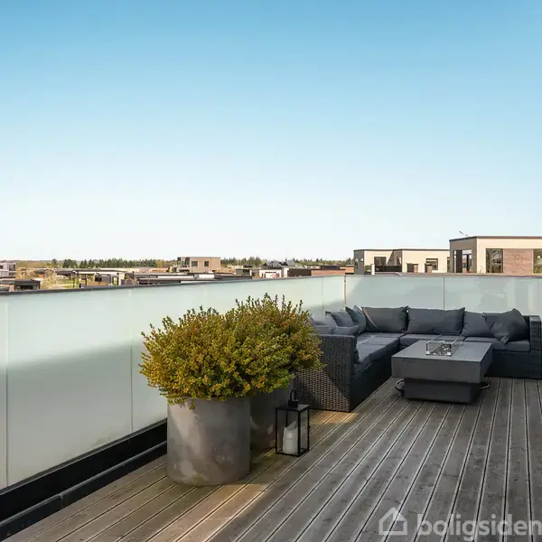 Terrasse med trædæk indeholder en grå sofa og et sofabord. En stor plante i krukke står ved glasrækværket, med udsigt til et boligområde og blå himmel i baggrunden.