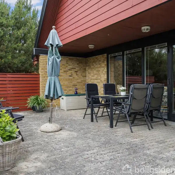 Terrassebord med stole står på en stenterrasse. En parasol er placeret ved siden af bordet. Området er omgivet af en rød trævæg og grøn beplantning.