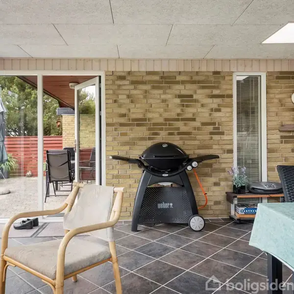 En gasgrill står på en terrasse med flisegulv, omgivet af stole og et bord med dug. En rød murstensvæg og planterammer ses i baggrunden. En dansk flagstang står ved siden af.