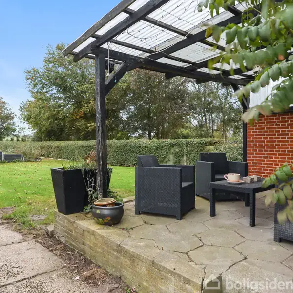 Terrasse med sorte havemøbler under en pergola, placeret ved siden af et rødt murstenshus. Omgivelserne består af et græsområde med træer og buske i baggrunden.