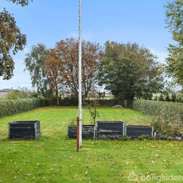 Flagstang står i en have omkranset af grønt græs og buske, med tre planterkasser foran. I baggrunden ses træer og en lys himmel. Tekst: "Boligsiden" ignoreret.
