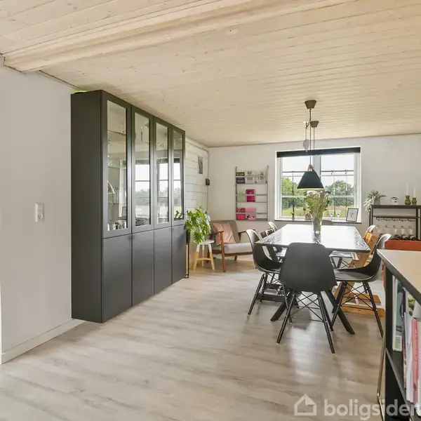 Stue med lang sort spisebord, omgivet af stole. Glasvitrineskab til venstre, lyse trægulve. Stor vindue i baggrunden. Grønne planter og hylder med dekorative genstande i rummet.