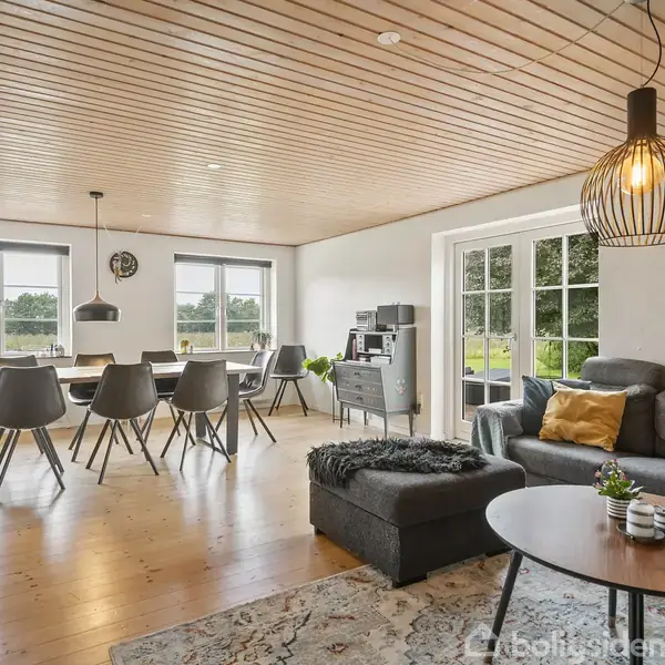 Stue med gråt hjørnesofa, sofabord og stor spisebord med seks stole. Rummet har lysindfald fra store vinduer og en dør til haven. Mønstret tæppe på trægulv.