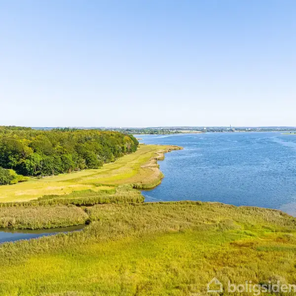 Et øde kystområde, hvor en grøn skov grænser op til en lys blå sø. Det omkringliggende område har en blanding af græsarealer og buske under en klar blå himmel.