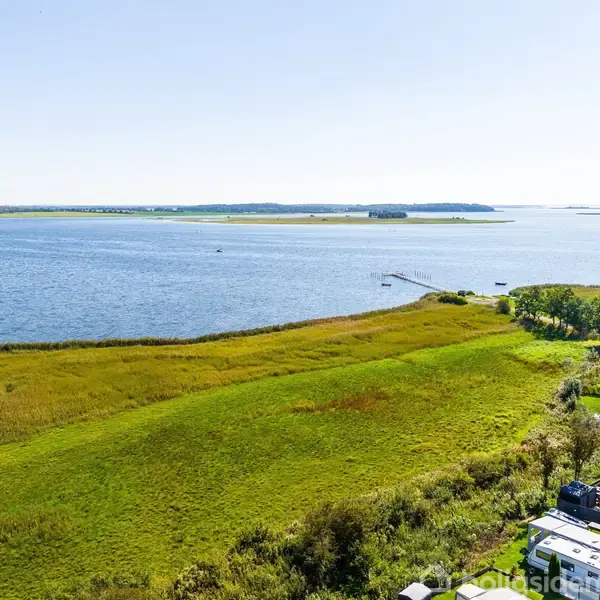 Græsmark med campingvogne står stille nær vandet, omgivet af frodig vegetation og udsigt over en bred strækning af vand mod horisonten.