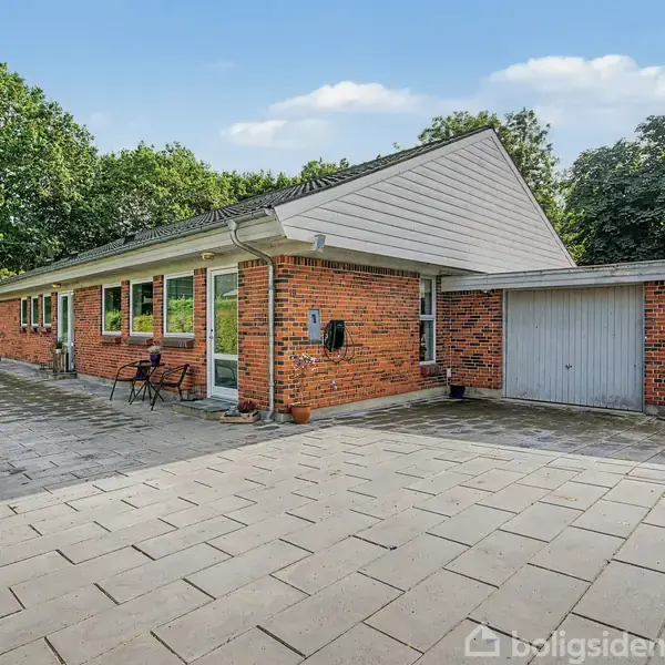 Et rødt murstenshus med garage står på en flisebelagt gårdsplads. Omkring huset er der frodig vegetation med grønne træer. Himmel med få hvide skyer overskygger stedet.