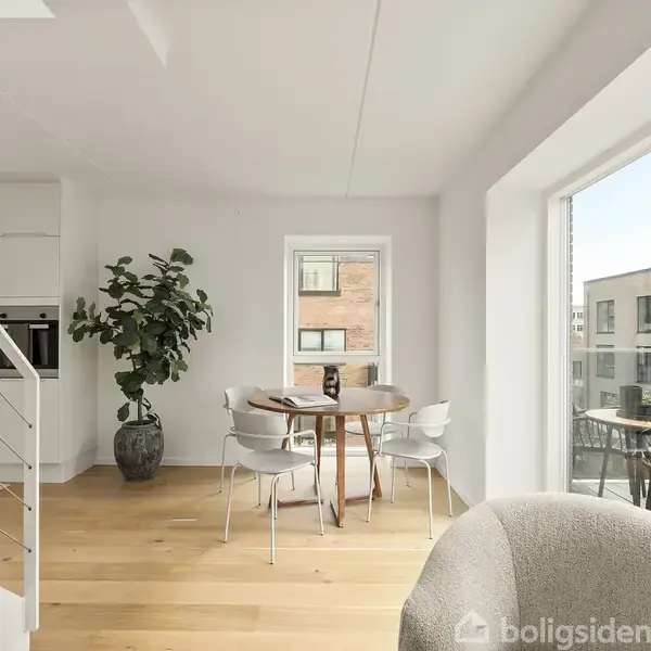 Et rundt spisebord med stole står ved et vindue i en lys stue. Der er en plante i hjørnet og udsigt til andre bygninger gennem de store vinduer.