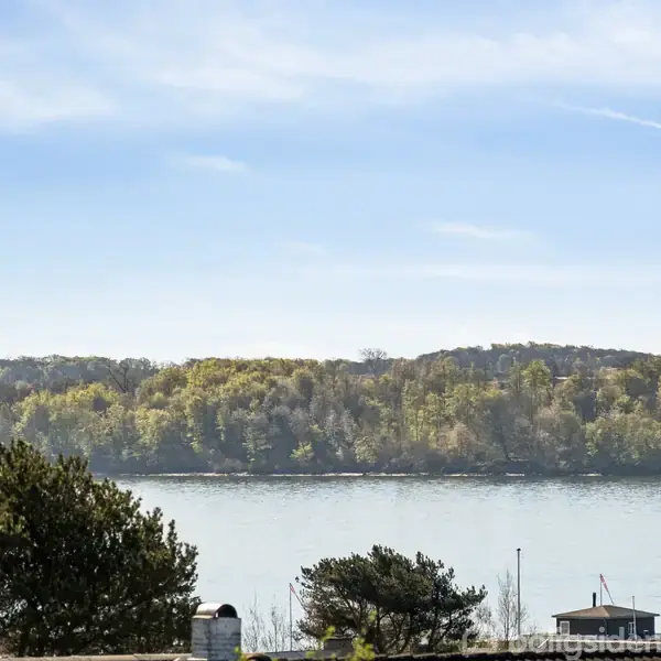 En grussti fører ned til en lille bungalow omringet af frodig beplantning. I baggrunden ses en sø eller fjord med en skovklædt kystlinje på den modsatte side.