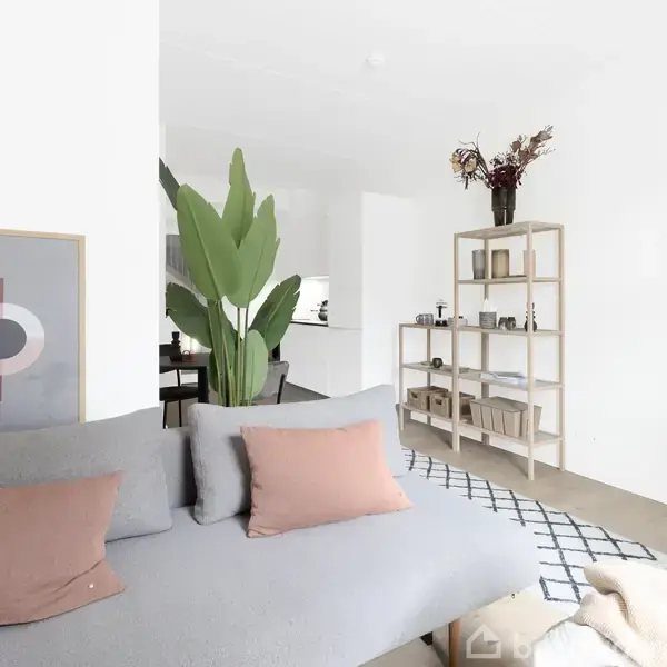 Grå sofa med brune puder står i en lys stue med abstrakt kunst på væggen. Plante og reol med bøger og dekorationer skaber en hyggelig atmosfære.