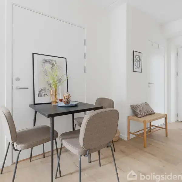 Et lille spisebord med fire stole står i en moderne, lys stue. På bordet er en vase med blomster og dekorationer. En bænk med en pude ses til højre.