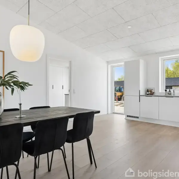 Et moderne køkken-alrum med et langt spisebord og sorte stole. En stor lampe hænger over bordet. Køkkenet har hvide skabe, og rummet er lyst med et vindue og dør til udsiden.
