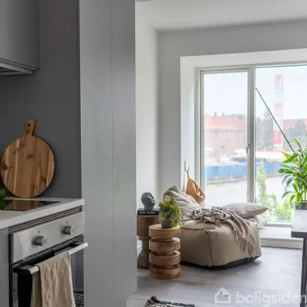 Et køkken-opholdsrum med et moderne køkken til venstre, dekoreret med skærebrætter og krydderier. Foran glasdøre til højre står en stol ved siden af et bord og grønne planter.