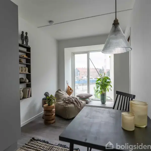 Stue med en sort spisebord, nogle stearinlys, og hængende lampe. En stol står ved bordet. I baggrunden er en lænestol ved et stort vindue med en plante ved siden af.
