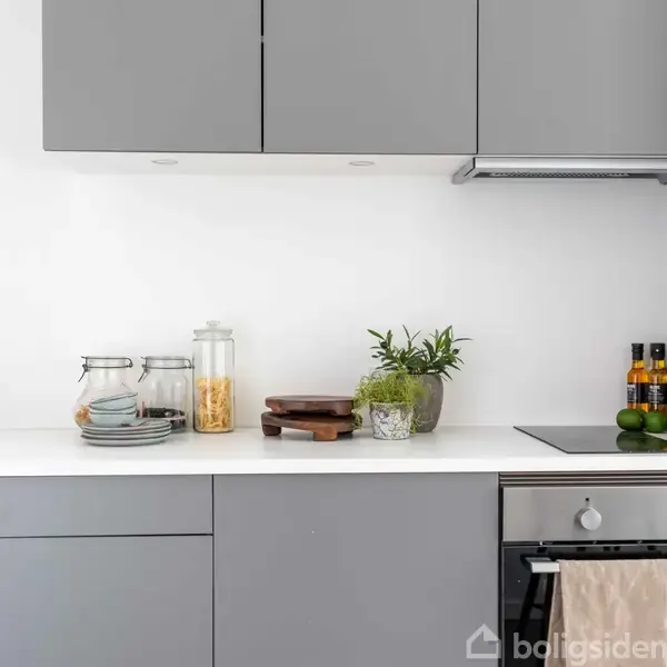Gråt køkken med indbyggede skabe. Tallerkener og glasbeholdere står ved vasken, mens plante, skærebræt, krydderier og limefrugt er på bordpladen ved ovnen. Lyst og moderne miljø.