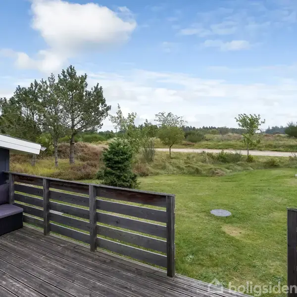 Terrasse med træbrædder og sofa vender ud mod en stor græsplæne. Omkringliggende træer ses under en blå himmel med få skyer.