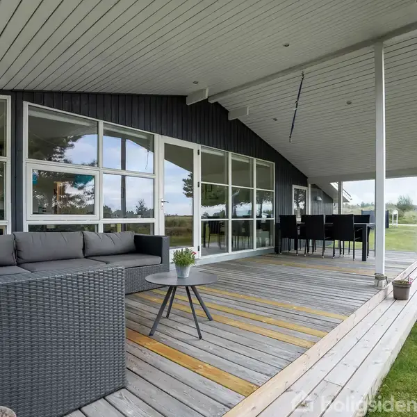 En udendørs terrasse med en grå sofa og et lille bord, der står under et overdækket tag. I baggrunden er der et spisebord med stole. Grøn græsplæne omgiver området.