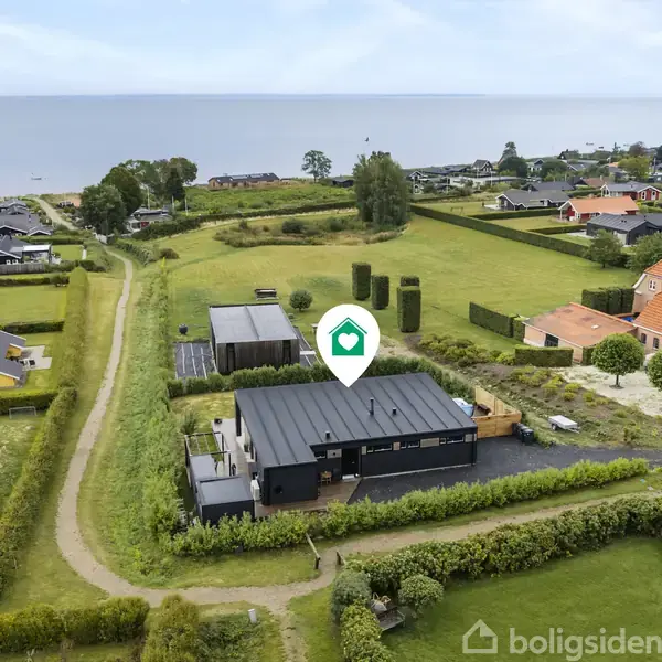 Et hus med fladt tag står omgivet af grønne haver og stier nær kysten. Baggrunden viser flere huse og en rolig havudsigt. Boligikonet svæver over ejendommen.