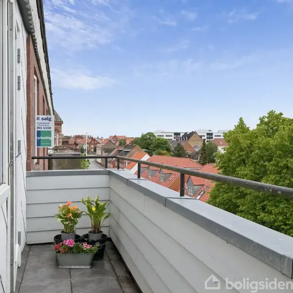 En altan med blomster i potter placeret langs rækværket. Altanen giver udsigt over en by med røde tegltage og grønne træer under en blå himmel.