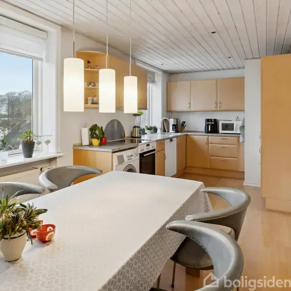 Køkken med spisebord dækket med lys dug og planter. Køkkenskabe og apparater ses i baggrunden, med tre hængelamper ovenover. Vindueskarm med planter giver udsyn til træer udenfor.