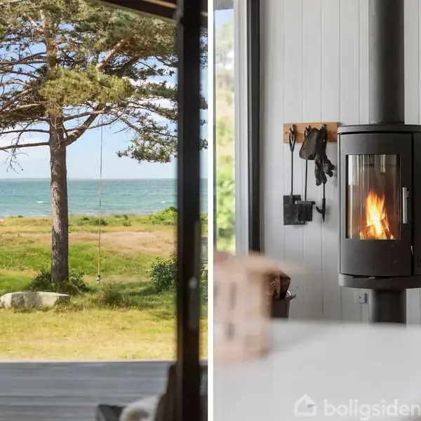 En pejseovn brænder lystigt i et moderne interiør. Til venstre en udsigt over græsplæne og træer mod havet, ses gennem en åben dør.