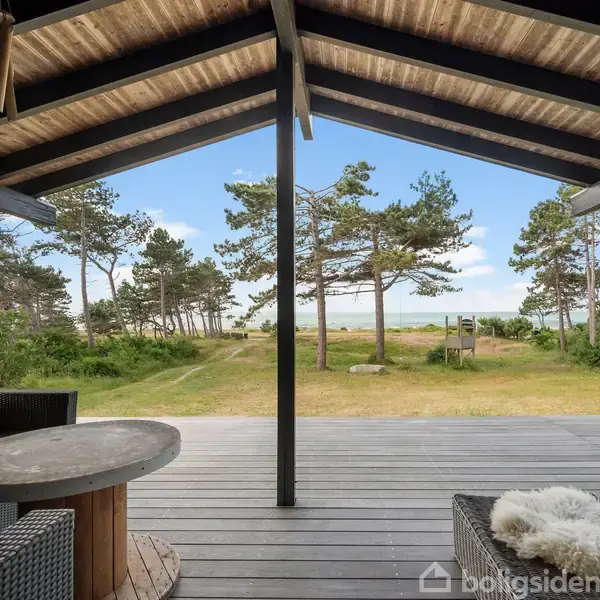 Træterrasse med sofa og bord under overdækning, med udsigt over græsplæne og træer mod havet i baggrunden. Himmel er klar med spredte skyer.