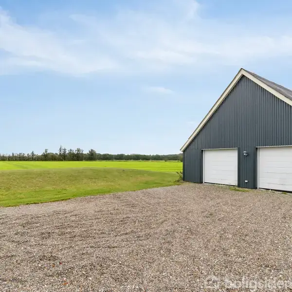 En grå garagebygning med to hvide porte står på en grusplads. I baggrunden kan ses en stor, grøn mark under en klar, blå himmel.