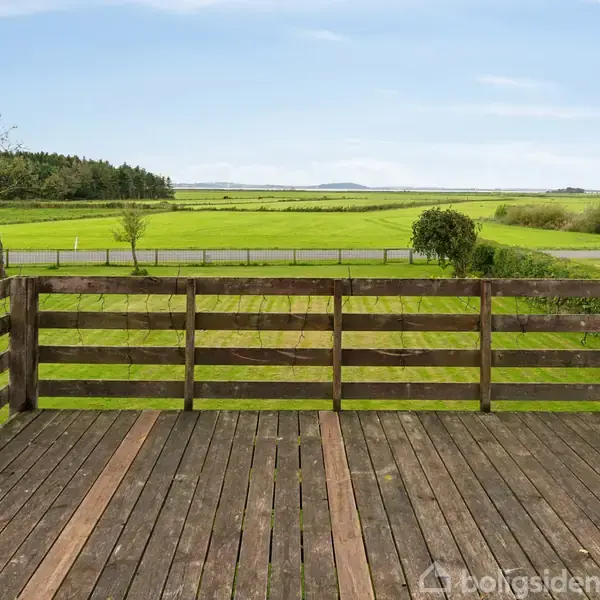 Træterrasse med udsigt over en grøn mark, indrammet af hegn. I horisonten ses et hus og træer langs kanten af marken. Himmel er blå med få skyer.