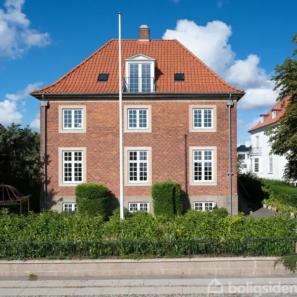 Et rødt murstenshus med rødt tegltag står i en grøn have omgivet af buske og træer. En flagstang er foran huset under en blå himmel med hvide skyer.