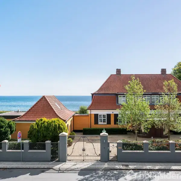 Et rødt taghus med grønne omgivelser står foran en blå havudsigt. Huset har store vinduer og en velplejet have adskilt fra vejen med et klassisk hegn og port.