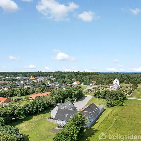 Spiseborde og stole står i en lys spisestue med hængelamper og vinduer mod en grøn have. Et køleskab i venstre side indeholder drikkevarer.