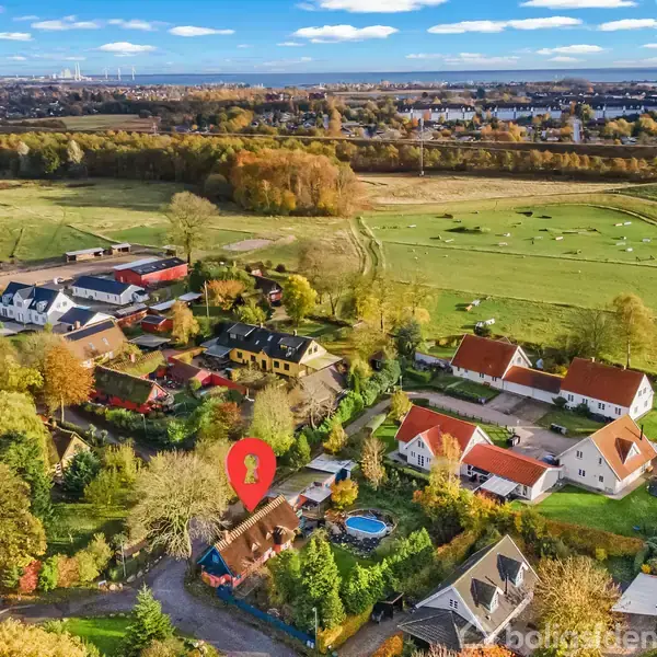 Luftfoto viser en landsby med spredte huse med røde tage omgivet af marker og træer. En rød markør angiver et specifikt hus. Bebyggelsen ligger i et landligt område.