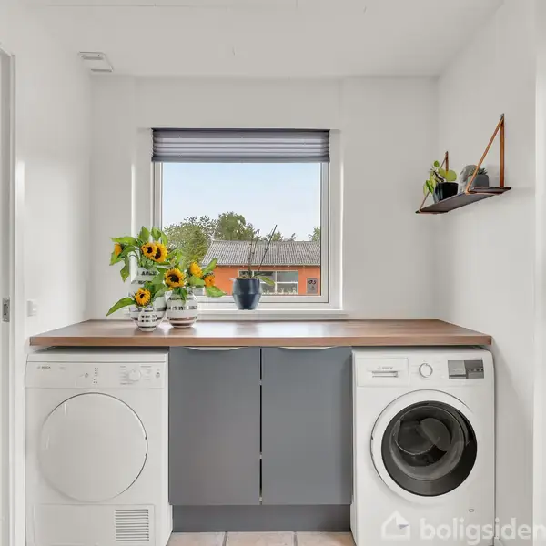 Vaskemaskine og tørretumbler står under en træbordplade med en vase med blomster. Rummet har hvide vægge, et vindue med persienner, og nogle planter på en hylde.