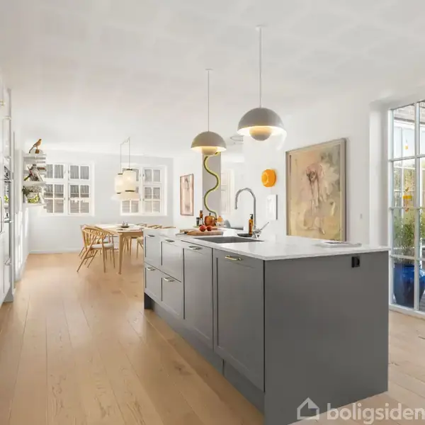 Et moderne køkken med en grå ø og to pendellamper. I baggrunden er et spisebord med stole. Store glasdøre åbner ud til terrassen. Malerier pryder væggene.