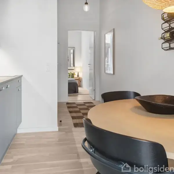 En rund spisebord med sorte stole står i et moderne køkkenalrum. Ved siden af står der en vase med blomster. I baggrunden ses en dør mod et andet rum.